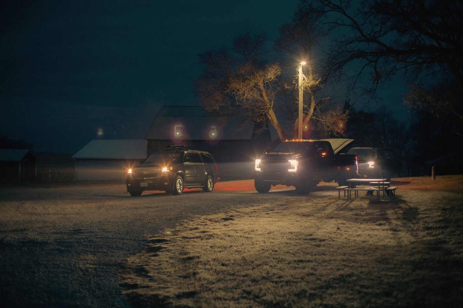Two pickups with headlights on beside a barn under a single pole light at night
