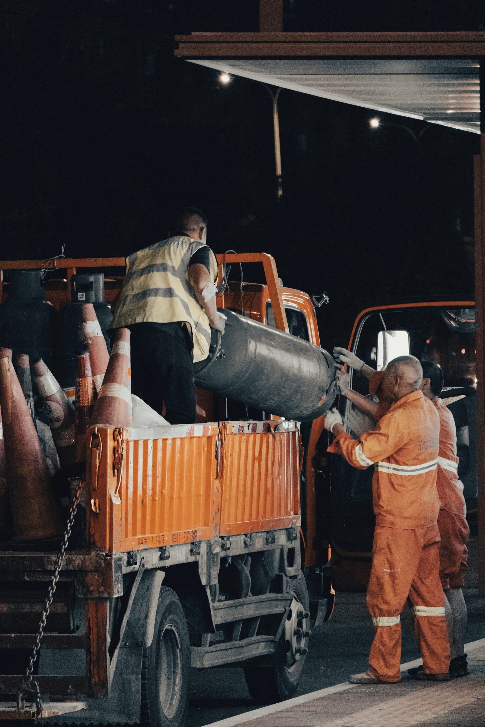 Two road workers in orange reflective coveralls loading equipment onto a truck at night