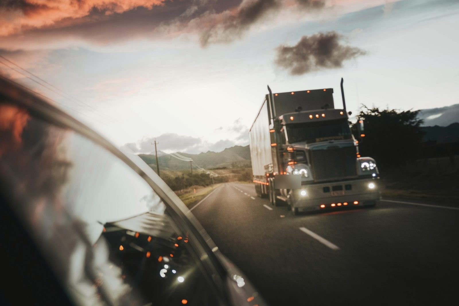 A semi truck with marker lights on, seen past a car mirror at dusk