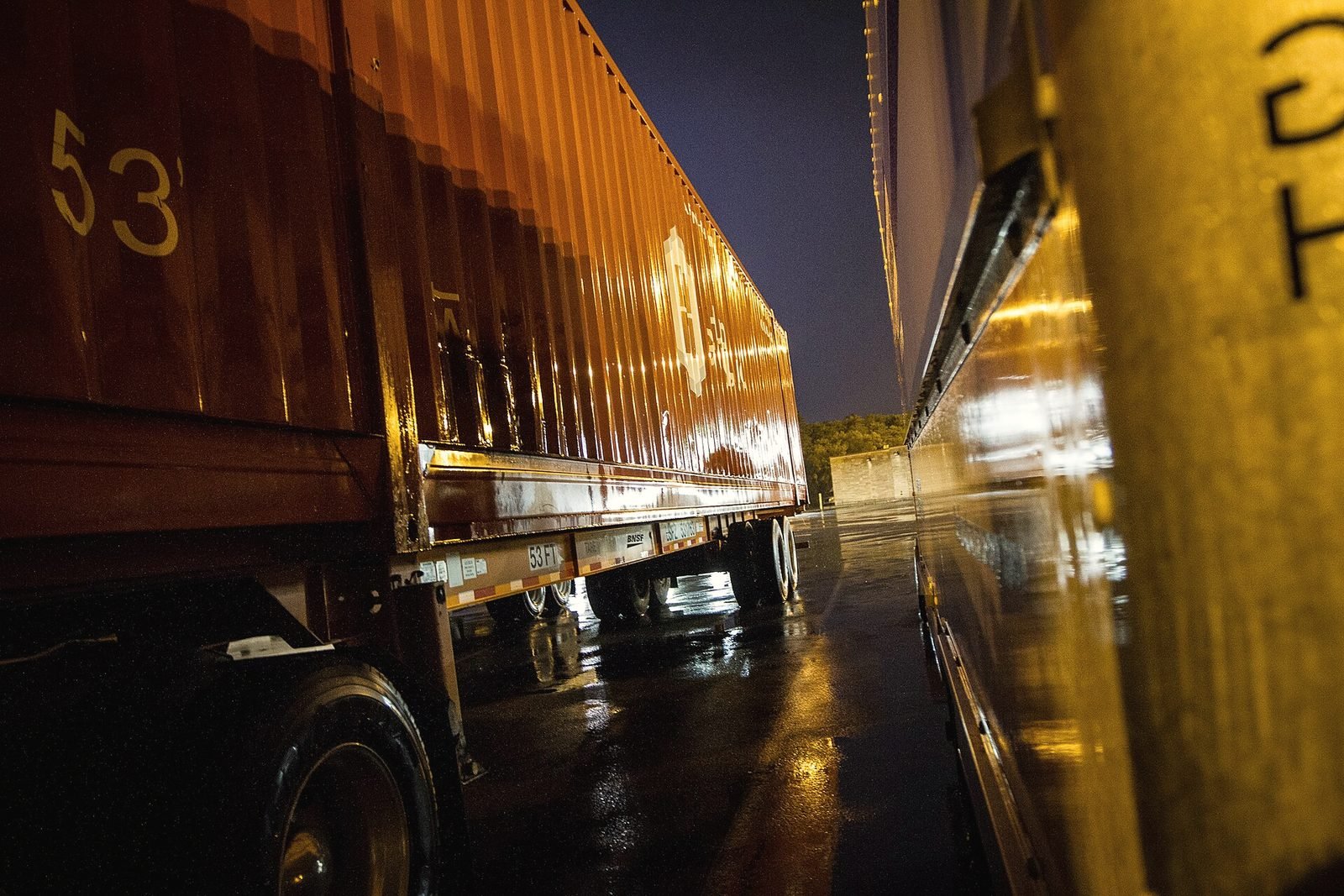 Two semi trailers parked side by side in the rain at night, lit amber by truck-stop lamps